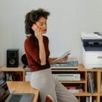 Stampante multifunzione a colori Xerox® C325 in un ambiente di ufficio domestico Una donna seduta alla scrivania mentre fa una chiamata accanto a una stampante Xerox® C325