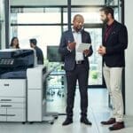 Due uomini in un ufficio discutono di qualcosa su un tablet accanto a una stampante multifunzione a colori Xerox® AltaLink® serie C8200 Due uomini in un ufficio discutono di qualcosa su un tablet accanto a una stampante multifunzione a colori Xerox® AltaLink® serie C8200